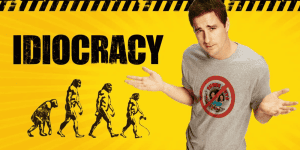 Man wearing a grey shirt with "Idiocracy" logo, gesturing with hands, yellow background.
