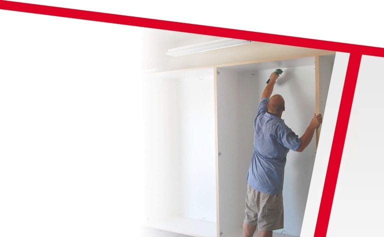 A professional interior renovation photograph showing a middle-aged man in a light blue short-sleeve shirt and khaki shorts installing a ceiling light fixture. The person is reaching up with both hands while standing on a white-painted wall. The image is framed by a bold red diagonal line in the top left corner, creating a modern, dynamic composition. The background is a pristine white room with a fluorescent light fixture visible on the ceiling. The lighting in the image creates subtle shadows and highlights the clean, minimalist space. The photograph is cropped to emphasize the action of installation work, with the red line serving as a design element that draws attention to the central figure and their task.
