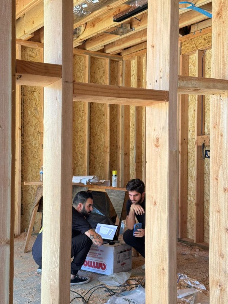 A construction site interior showing two caucasian men working inside a wooden-framed building. The walls and ceiling are made of exposed light-colored wooden studs and beams. The floor is unfinished with sawdust and construction debris scattered about. A white OSB panel covers the wall surfaces. The workers are crouched down, examining something on a tablet device. A white cardboard box with red text is visible on the ground. The lighting is natural, coming from an unseen source, creating subtle shadows through the wooden framework. The ceiling shows exposed black electrical wiring and insulation materials. The wooden beams are Douglas fir with a natural golden color. The image is taken from a side angle, showing the structural framework of the building.