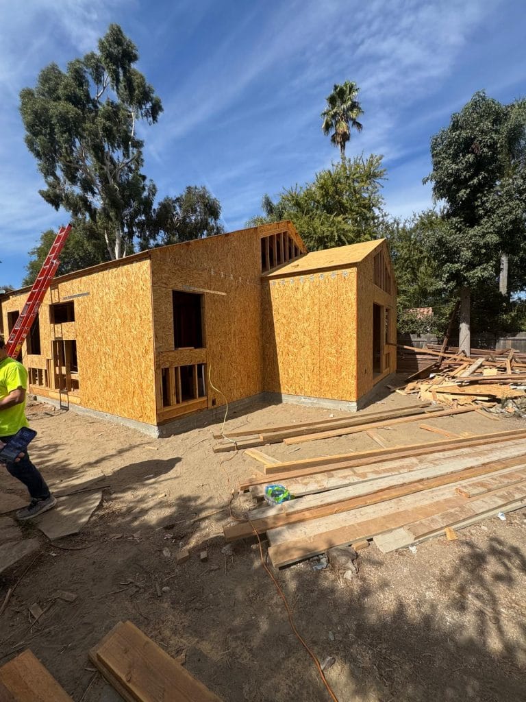 A construction site photograph of a partially built house with OSB (oriented strand board) exterior walls. The house has a modern design with two distinct sections - a rectangular main structure and a detached garage-style extension. The OSB panels are golden-yellow in color and cover the exterior walls. A red extension ladder leans against the left side of the house. The ground is unpaved dirt with scattered construction materials and lumber. In the background, there are mature eucalyptus trees with dark green foliage and a tall palm tree. The sky is bright blue with wispy white clouds. A construction worker in a yellow safety vest is partially visible on the left edge of the frame. The scene is photographed during daytime with natural sunlight casting shadows from the trees onto the construction site.