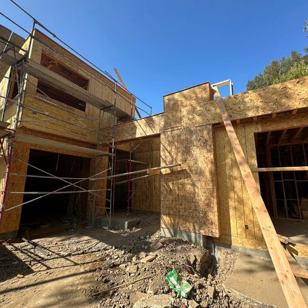 A construction site photograph of a modern house under development. The structure features exposed wooden OSB sheathing and metal scaffolding. The building has multiple levels with large garage openings on the ground floor. The scaffolding is made of silver metal poles and cross-braces. The ground is unpaved with dirt and gravel scattered across the construction site. The sky is a bright, clear blue. Green foliage from trees is visible at the top right corner of the frame. The architecture has clean lines with angular roof angles and large window openings. The wooden framing creates a grid-like pattern across the exterior walls. Construction materials and debris are on the ground, including a green plastic item partially visible in the frame. The lighting is from direct sunlight, creating strong shadows and highlighting the natural wood grain of the OSB panels.