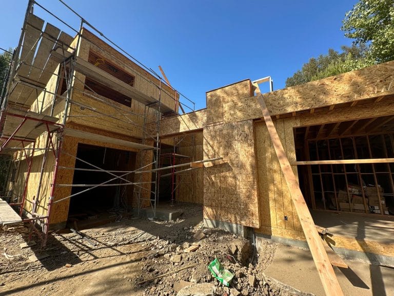 A construction site photograph of a modern house under development. The structure features exposed wooden OSB sheathing and metal scaffolding. The building has multiple levels with large garage openings on the ground floor. The scaffolding is made of silver metal poles and cross-braces. The ground is unpaved with dirt and gravel scattered across the construction site. The sky is a bright, clear blue. Green foliage from trees is visible at the top right corner of the frame. The architecture has clean lines with angular roof angles and large window openings. The wooden framing creates a grid-like pattern across the exterior walls. Construction materials and debris are on the ground, including a green plastic item partially visible in the frame. The lighting is from direct sunlight, creating strong shadows and highlighting the natural wood grain of the OSB panels.
