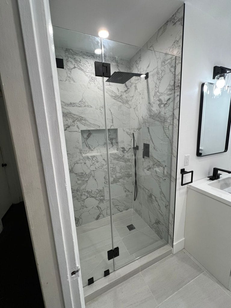 A modern, luxurious bathroom interior with a frameless glass shower enclosure featuring white and gray marble-patterned tile walls. The shower has a large square rainfall showerhead mounted on a black wall bracket and a handheld shower attachment. The floor of the shower is finished with large-format white tiles. The bathroom has white walls outside the shower area and a white floating vanity with a rectangular black-framed mirror mounted above it. The vanity has a white countertop and black faucet. The lighting includes recessed ceiling lights and a wall-mounted sconce with frosted glass globes. The marble wall tiles have dramatic gray veining patterns throughout the white surface. The shower door features black hardware and hinges. The floor outside the shower area matches the shower floor tiles. The overall color scheme is monochromatic with white, black, and gray tones, creating a contemporary and sophisticated aesthetic.