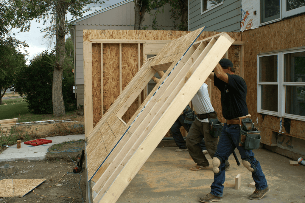 A construction scene showing three caucasian male workers installing a large wooden wall panel. The workers are wearing blue jeans, work boots, and tool belts. The wall panel is made of oriented strand board (OSB) and is being lifted into place against a partially finished exterior wall. The exterior wall has exposed OSB panels and a white-framed window. The background shows a light gray house with vinyl siding and trees with green foliage. The ground is dirt and gravel, with construction debris including a red tool on the ground. The lighting shows it's daytime, with natural outdoor lighting casting soft shadows. The workers are positioned at the base of the wall panel, with one worker in a black shirt actively participating in the installation while the others are assisting.