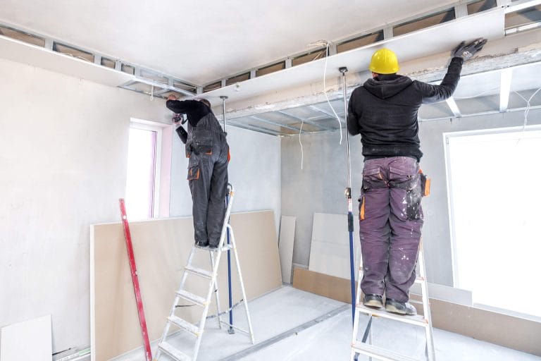 A construction interior renovation scene with two caucasian men working on a ceiling. The workers are standing on two separate metal ladders - one white aluminum ladder and one blue ladder. The man on the left wears a black work uniform with orange accents and is reaching up towards the ceiling. The man on the right wears purple work pants and a yellow hard hat, also reaching towards the ceiling. The room has white walls and is partially covered with beige floor protection. The ceiling is exposed with metal framework visible. There is a window on the left wall with a pink frame. Various construction tools and equipment are scattered around the room, including a red tool on the floor. The lighting is bright and even, creating a clean, well-lit workspace atmosphere. The image shows the room in a state of renovation, with drywall panels visible on the walls.