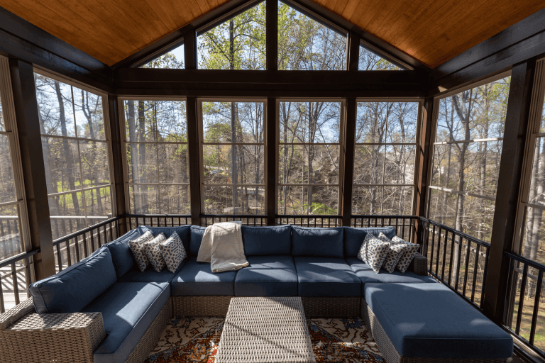 A modern screened-in porch with a high peaked ceiling and large windows. The space features a large navy blue L-shaped sectional sofa with matching throw pillows and a white throw blanket. A white woven coffee table sits in front of the sofa on a patterned area rug with red and beige tones. The porch has black metal railings and is surrounded by floor-to-ceiling windows that provide a view of bare trees and green foliage outside. The ceiling is finished with warm wood paneling, and natural sunlight streams through the windows, creating dappled shadows on the furniture. The flooring is made of light-colored tiles. The room has a clean, contemporary aesthetic with a neutral color palette dominated by blues, whites, and natural wood tones.