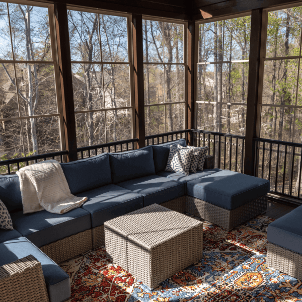 A modern screened-in porch with floor-to-ceiling windows and black metal railings. The space features a large L-shaped sectional sofa with navy blue cushions and wicker frame. The sofa is adorned with decorative pillows in gray and white geometric patterns. A large beige wicker ottoman serves as a coffee table in the center of the seating area. The floor is covered with a large area rug featuring intricate patterns in red, blue, and beige tones. The rug has traditional design elements with floral motifs. Through the windows, there is a view of bare winter trees and a wooded area. Natural sunlight streams through the windows, creating dappled shadows on the furniture and floor. The ceiling is dark-colored, and the overall color scheme consists of navy blue, beige, and natural wood tones. The space is well-lit with both natural and ambient lighting, creating a cozy indoor-outdoor living environment.
