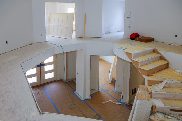 A high-angle interior construction photo of a modern home renovation. The space features a curved staircase with wooden treads and white risers on the right side. The floor is covered with brown cardboard protection material and blue painter's tape marking lines. The walls are painted white with exposed electrical outlets. A white paneled door is visible in the upper portion of the frame. The ceiling has exposed beams and is partially finished. Construction materials and debris are scattered throughout, including a red tool and cardboard boxes. The lighting is bright and even, creating clear visibility of all construction elements. The space has multiple levels and angles, with a distinctive architectural design featuring curved elements and open floor plans.