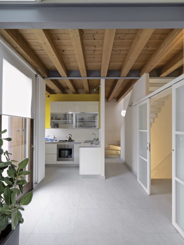 A modern minimalist kitchen interior with exposed wooden ceiling beams. The ceiling features natural pine wood beams with dark metal supports running parallel across the space. The kitchen has white cabinets with glass-fronted upper cabinets painted in a bright yellow. The lower cabinets and countertops are white with a stainless steel sink and built-in oven. The floor is covered in large format light grey tiles. A white sliding door is visible on the right side of the frame, leading to a staircase. A large green potted plant with broad leaves sits in the left corner of the room. Natural light streams in through a window on the left side, creating a bright, airy atmosphere. The walls are painted in a clean white color, and a simple white pendant light fixture hangs from the ceiling. The space has a clean, contemporary aesthetic with a warm wood accent from the ceiling beams.