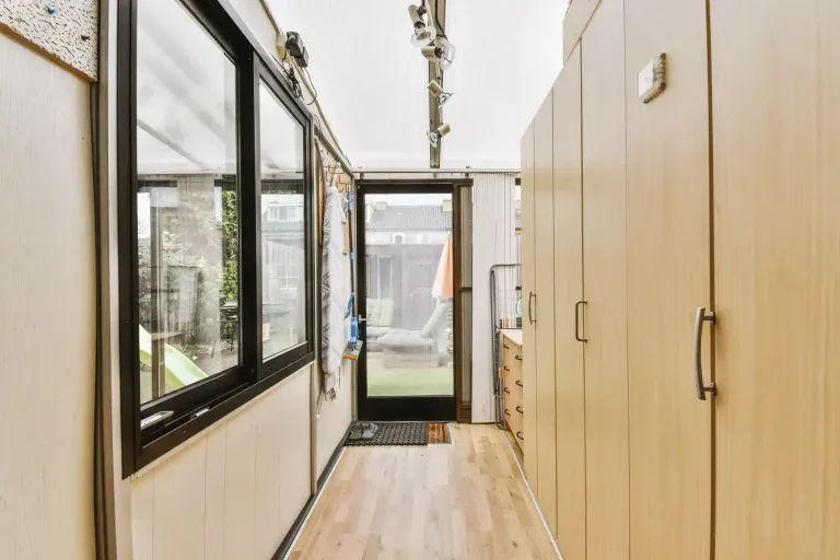 A modern interior hallway with light wooden flooring and cream-colored walls. The hallway features a row of large black-framed windows on the left side, allowing natural light to stream in and showing a view of greenery outside. The right wall has floor-to-ceiling light wood paneling with metal handles. A black metal door is at the end of the hallway, with a black doormat placed in front of it. Track lighting fixtures with multiple heads are mounted on the ceiling, directed towards the windows. The hallway has a clean, minimalist design with straight lines and a narrow perspective. The windows are divided into multiple panes with black frames, creating a grid-like pattern. The flooring has a natural wood grain pattern and is laid in a horizontal direction. The overall color scheme consists of warm neutrals, blacks, and natural wood tones.