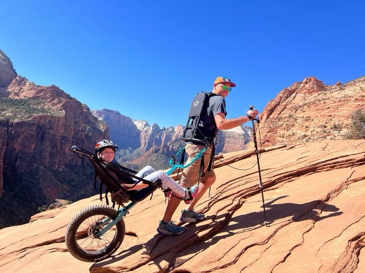 A man hikes up a rocky trail using trekking poles while pulling a child in an all-terrain wheelchair with one wheel.
