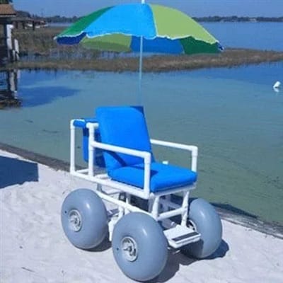 A beach wheelchair with large gray wheels, blue cushions, and a green-and-blue umbrella is positioned on sand near water.