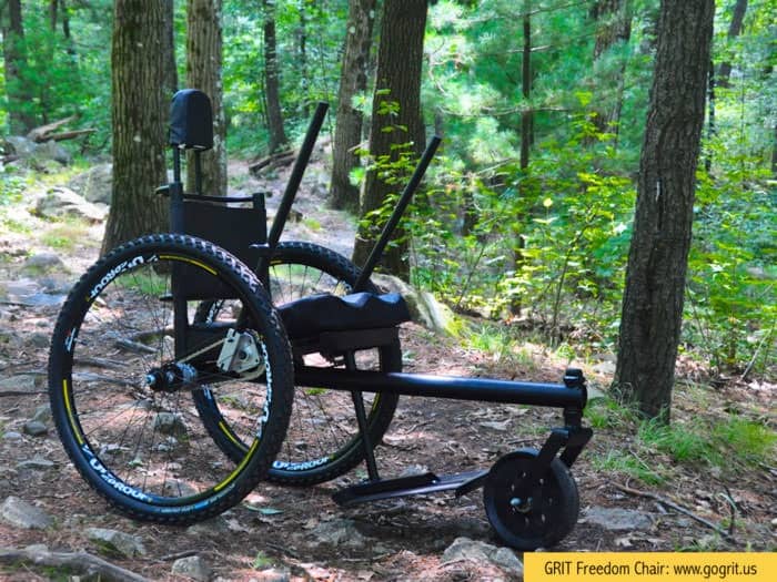 A rugged wheelchair with large off-road wheels and hand levers sits on a forest trail surrounded by trees.