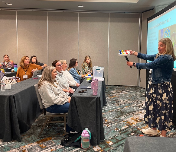 Lynn stands at the front of a classroom holding a tray with colorful objects, while a group of seated adults watches attentively. A presentation is projected on the screen.