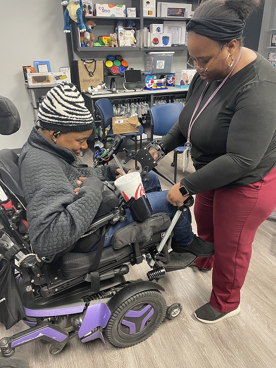 A woman in a wheelchair is assisted by another woman in an office setting.