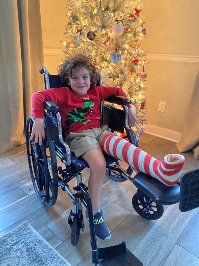 A young boy in a wheelchair with a red and white striped cast on his leg sits in front of a decorated Christmas tree, smiling at the camera.