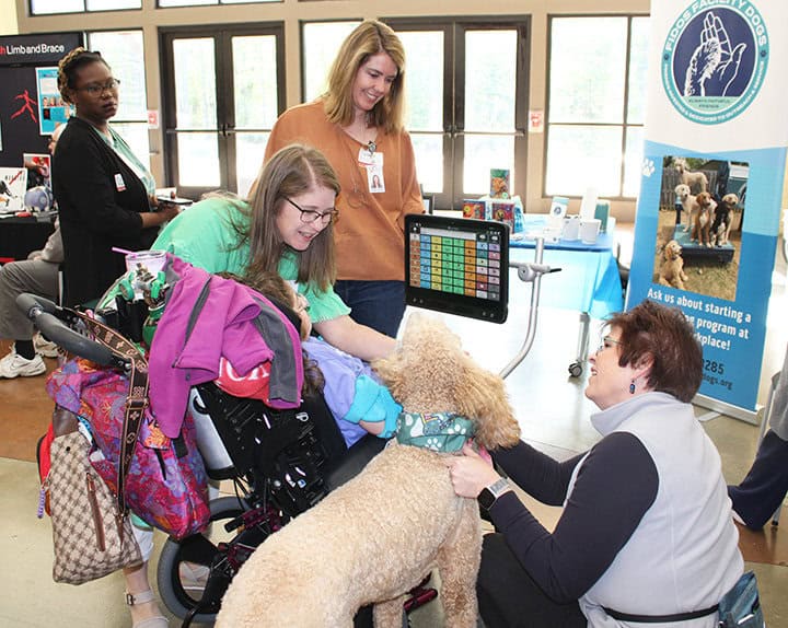 A woman pets a large curly-haired dog beside a person in a wheelchair while others look on.