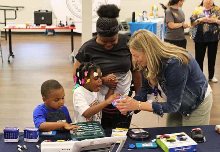 Lynn from APTAT shows two children and an adult a light-up device at a table with educational toys during an indoor event.