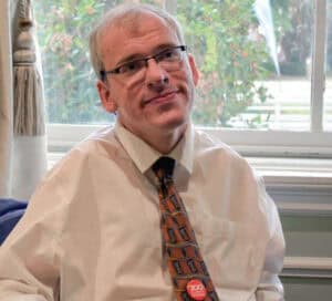 Eric Peebles has light hair and glasses, wearing a white shirt and patterned tie, sits indoors near a window with greenery visible outside.