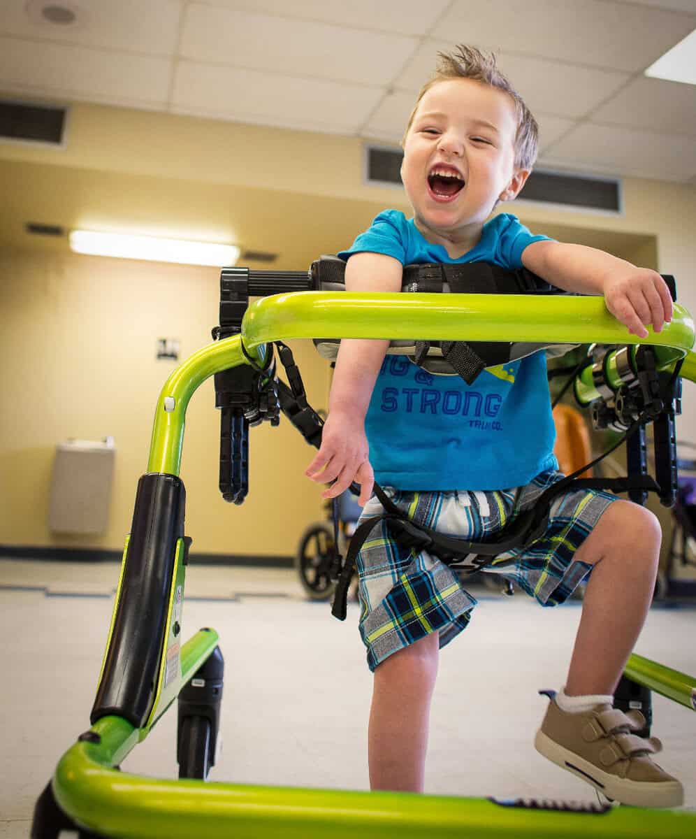 A young child with short hair smiles joyfully while using a bright green walker.