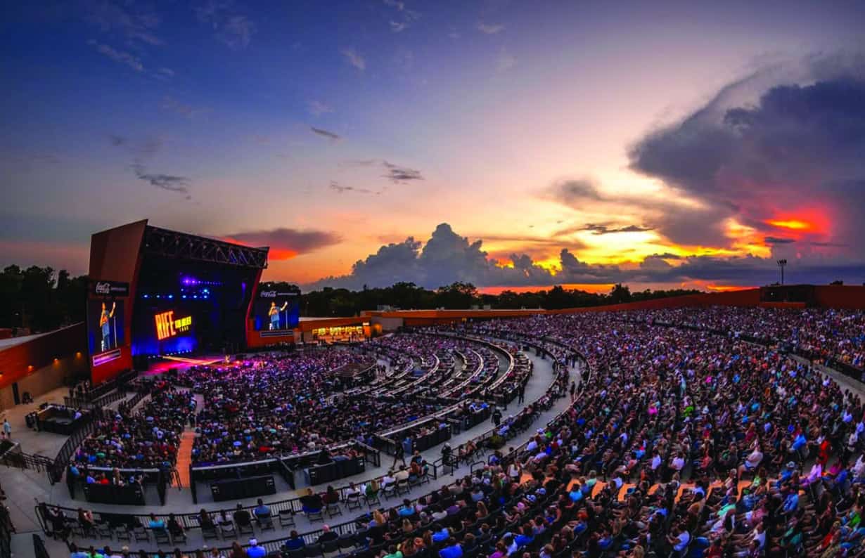 A large outdoor concert stadium filled with hundreds of people seated in circular rows watching a performer on stage during sunset. The sky is colorful with clouds and orange hues.