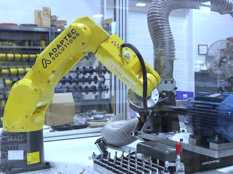 Yellow industrial robotic arm with Adaptec Solutions branding in a manufacturing setting.