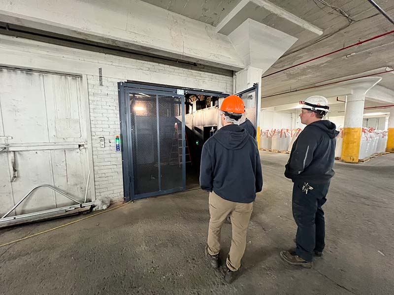 Two workers inspecting elevator machinery in parking garage.