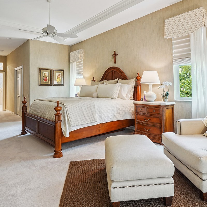 Bedroom with wooden bed frame, white bedding, nightstand with lamp, cross on wall, and large windows with white blinds and curtains, cozy interior, advance blinds & drapery.
