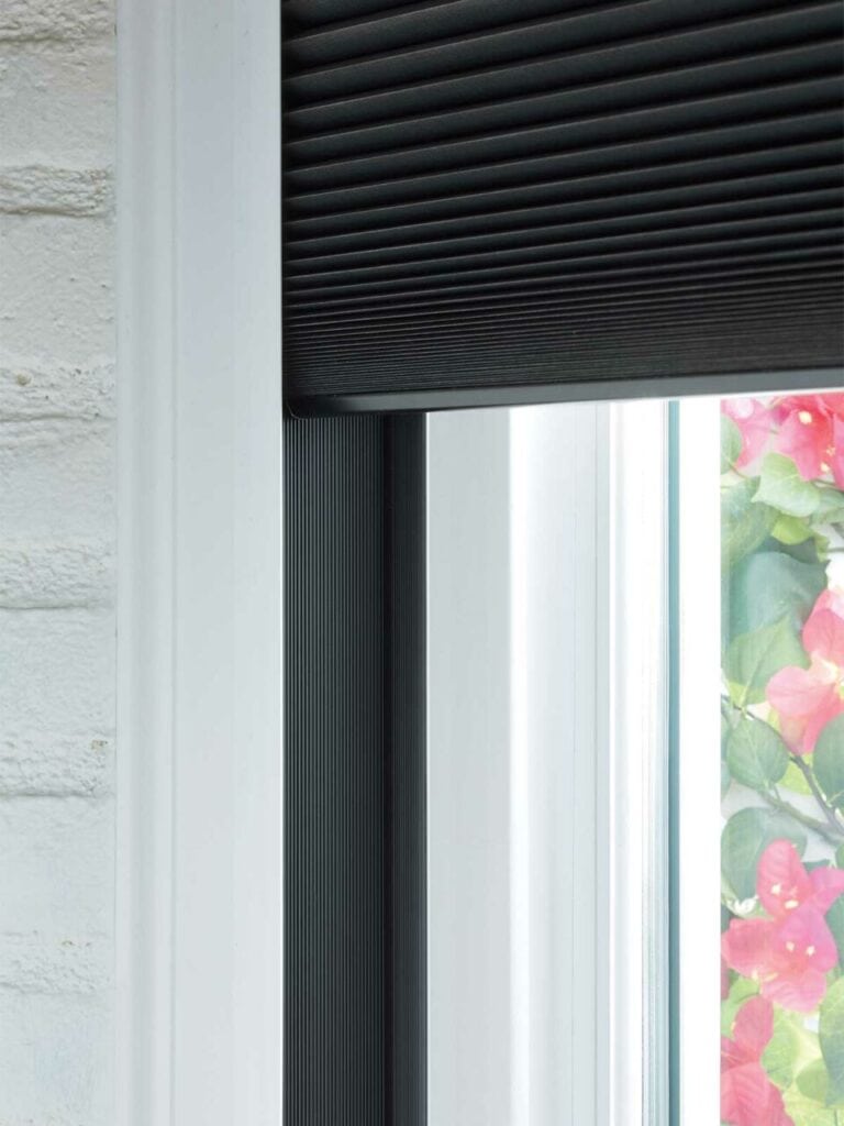 Black vertical window blind on a white frame with a glimpse of colorful flowers outside, offers stylish window covering solutions with premium blinds and drapery services from Advance Blinds & Drapery.