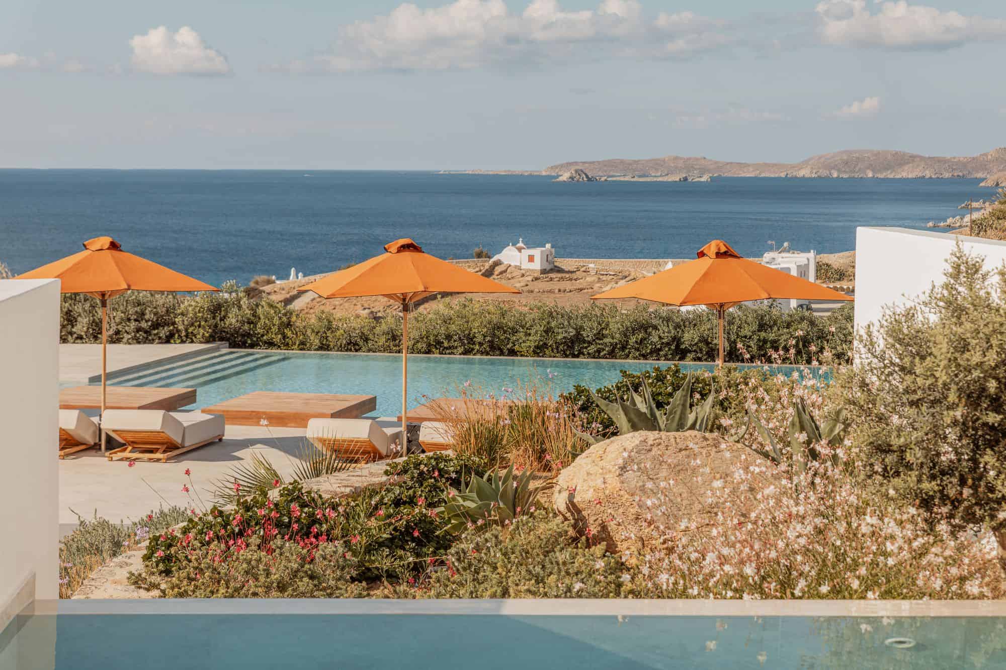 Stunning outdoor pool at Aelo Mykonos overlooking the Aegean Sea with vibrant orange umbrellas and lush landscaping.