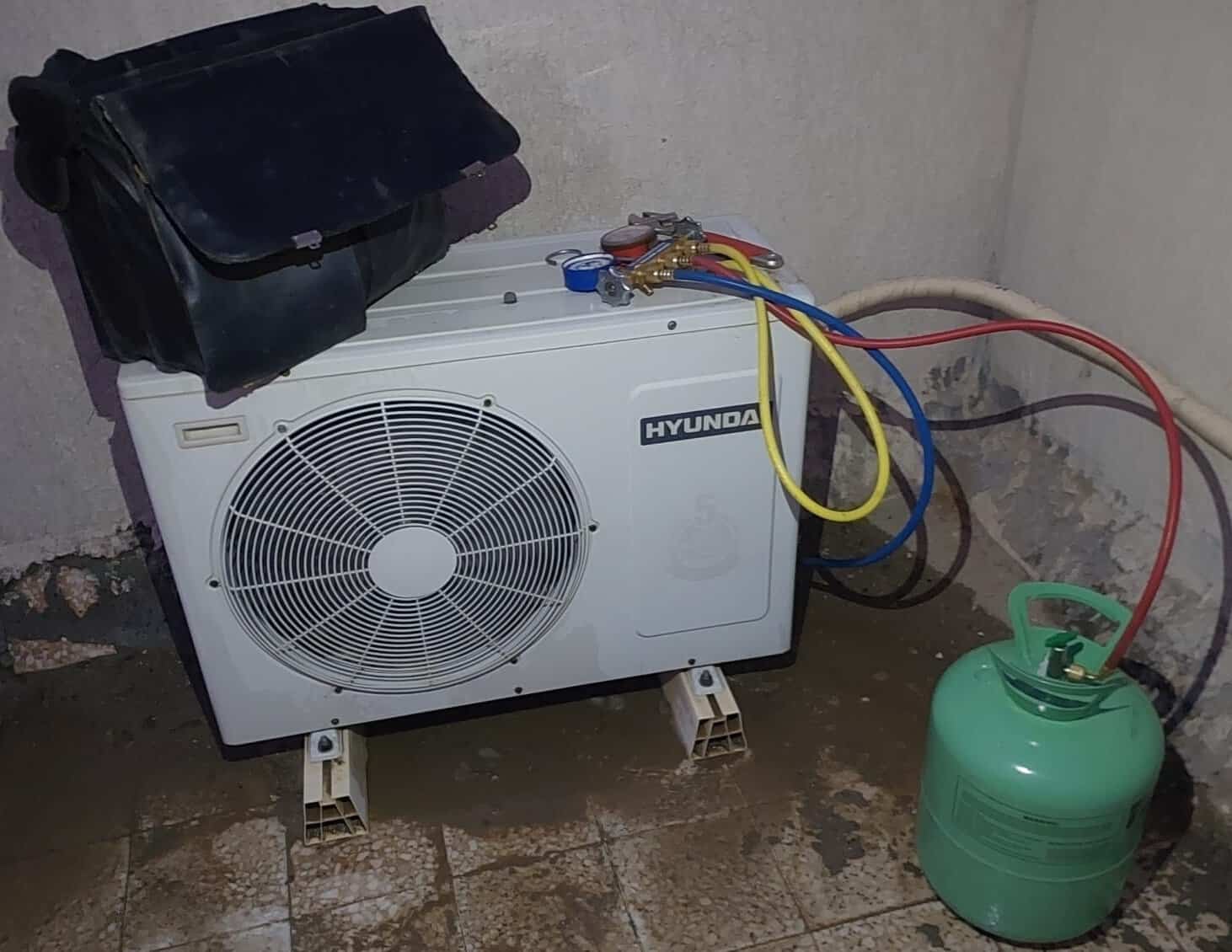 فني تكييف هندي بالكويت يقوم بصيانة وحدة تكييف هواء من نوع هيونداي مع أدوات ومجموعة غازات تبريد.