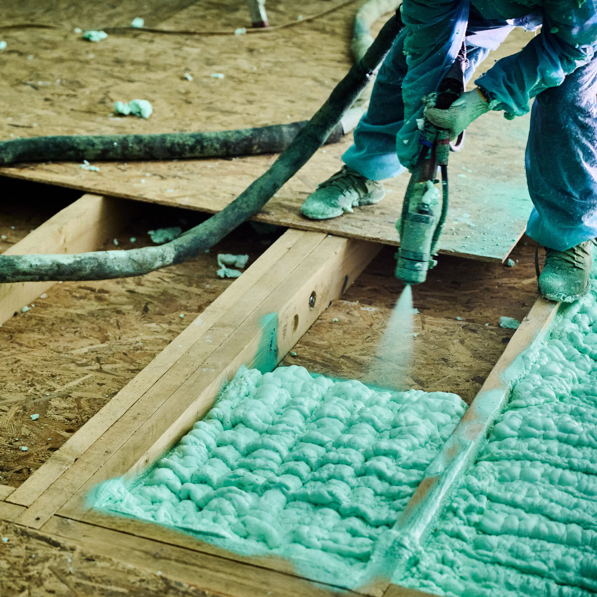 Worker applying green spray foam insulation to a wooden floor frame.