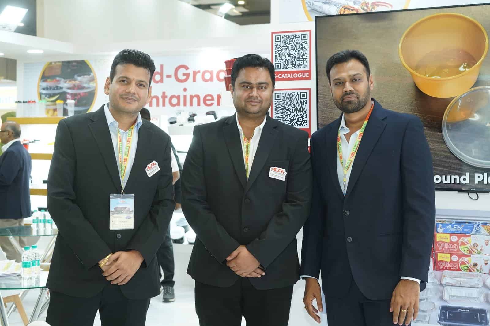 Three men in formal suits at Alfa Foil Containers booth at trade show.