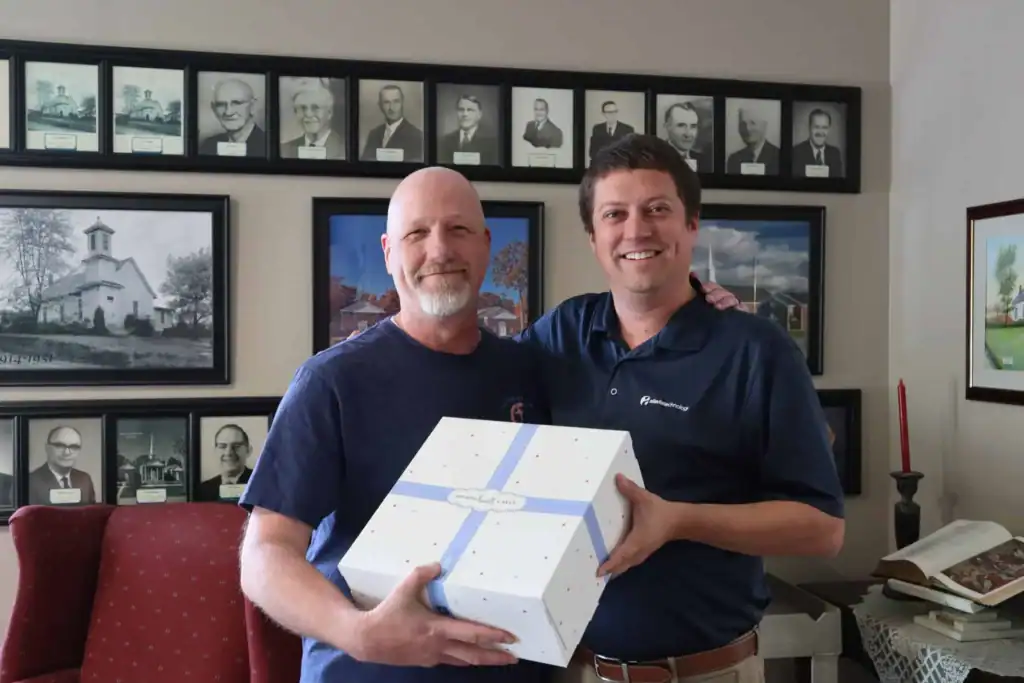 Two men smiling, holding a white gift box in an office setting.
