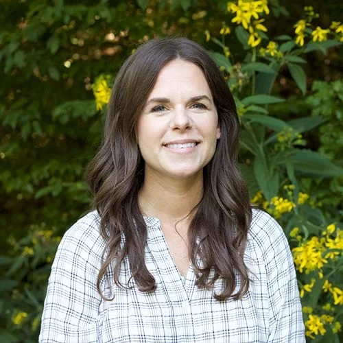 Smiling woman outdoors with greenery and yellow flowers in background.