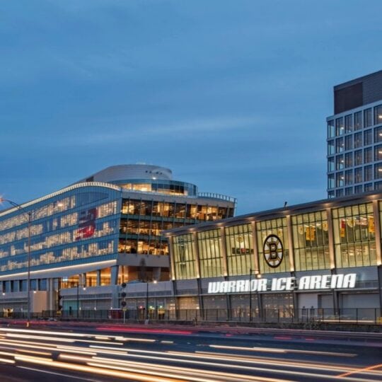 The image shows the Warrior Ice Arena building in an urban setting during dusk, with light trails fr.