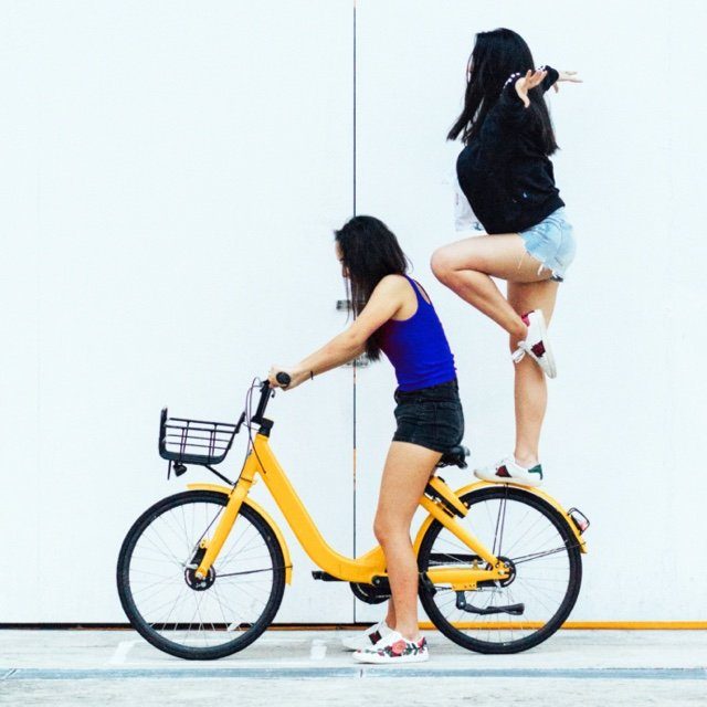 A woman in a blue top riding a yellow bike with another woman standing on the rear wheel, balancing.