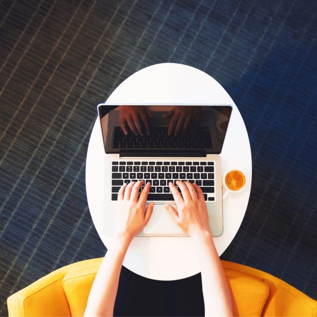 A person typing on a laptop at a white round table with a cup of coffee.