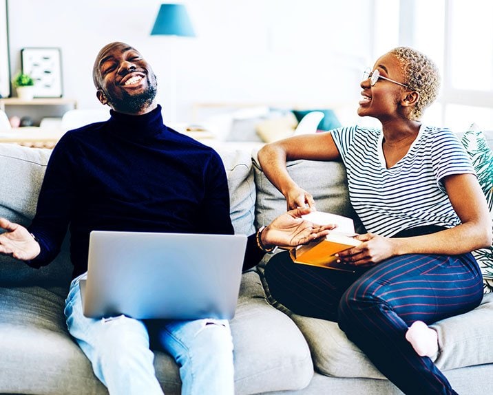 A smiling man and woman chatting on a sofa with a laptop and notebook.