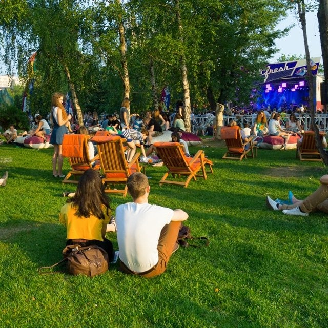 A lush green park with people sitting on grass and wooden chairs, enjoying an outdoor concert.
