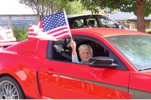 Summerset Resthome 11 Red sports car with elderly woman holding American flag in car window | vibrant red convertible sports car with smiling elderly woman waving American flag | elderly woman enjoying a ride in a red sports car, holding an American flag, celebrating American Heritage at Americarna | festive scene at Americarna showing a cheerful elderly woman in a red sports car waving an American flag during a cultural automobile event | relevance.