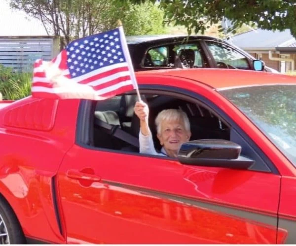 Red American car with elderly woman waving an American flag, celebrating at Americarna event in NZ, classic car, patriotic parade, vintage vehicle celebration.