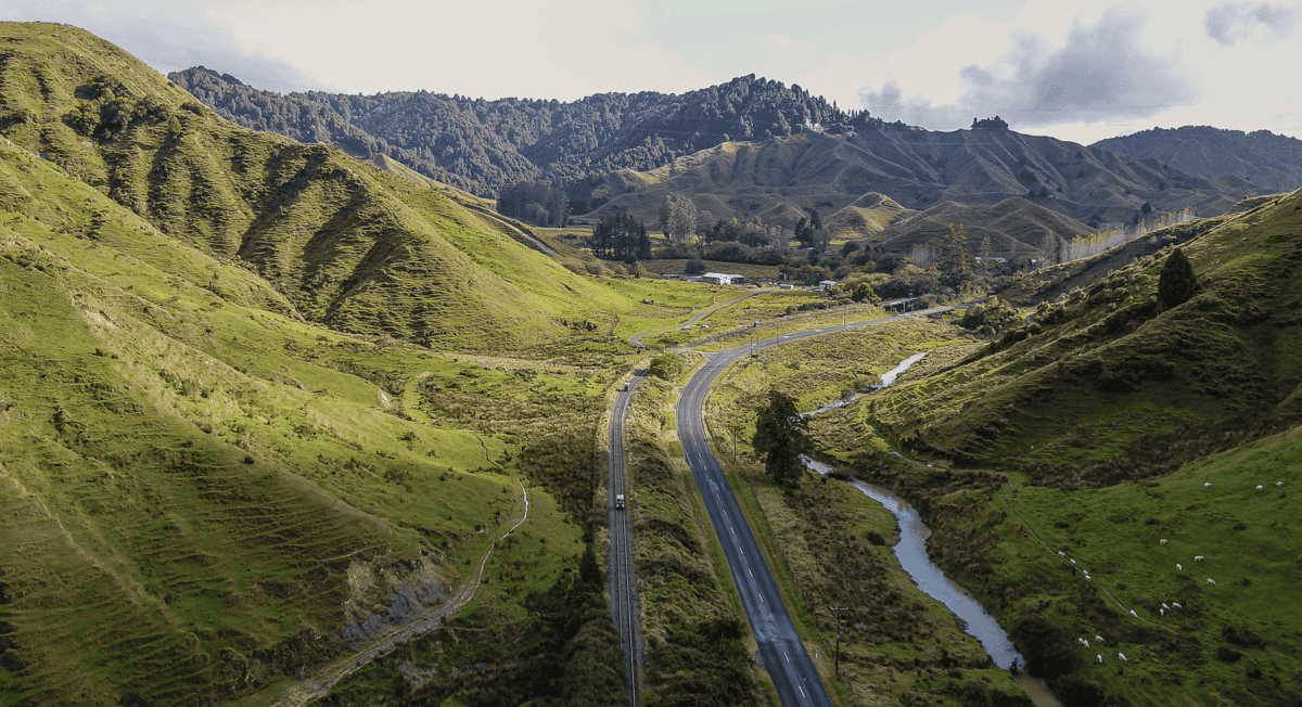 Melting green hills New Zealand scenery with winding road and river in the valley, lush landscape and mountain backdrop.