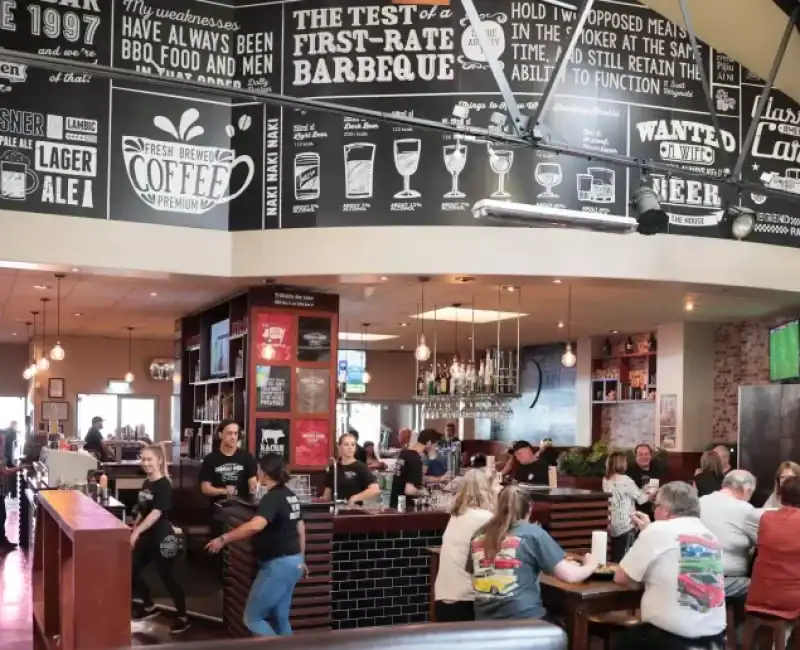Colourful bar and restaurant interior with patrons enjoying meals, bar staff preparing drinks, and multiple TV screens showing a sports game.