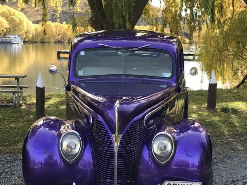 Classic American vintage car in shiny purple, parked by the river amidst autumn trees, perfect for car enthusiasts and collectors.