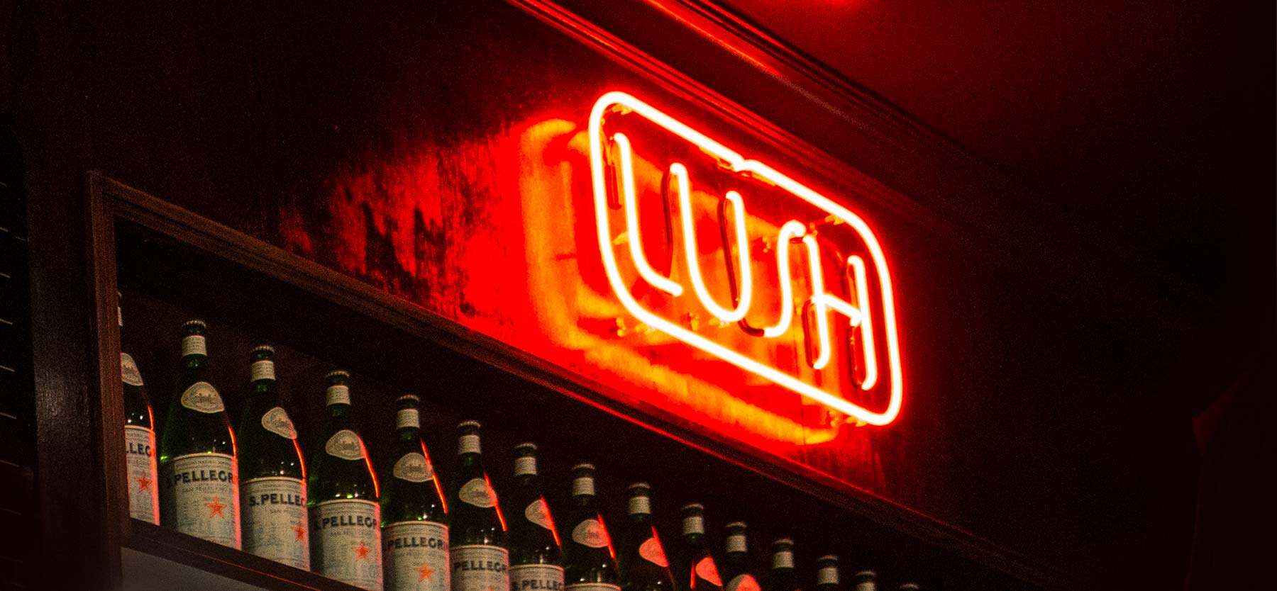 Neon "Open" sign lights up a vintage car show venue, with a row of beer bottles on display nearby.