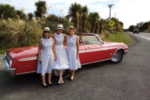 Vintage American car and three women in polka dot dresses and hats by roadside.