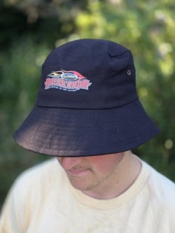 A person wearing a navy bucket hat featuring the Americana logo, outdoors with greenery in the background.