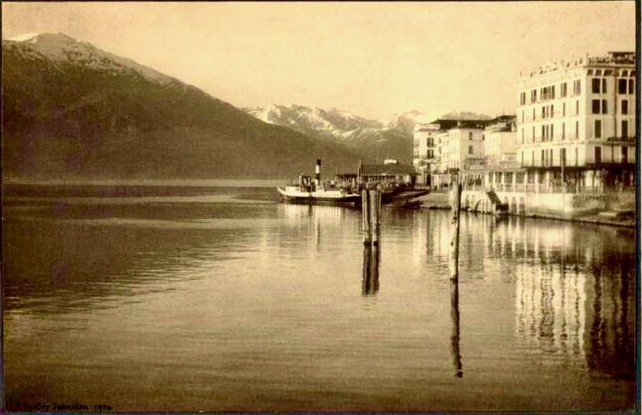 Vintage sepia photo of lakeside buildings and mountains in the background.