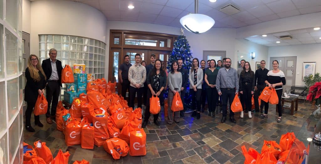 Porteurs de sacs cadeaux orange devant un sapin de Noël en équipe, distribution de Noël, solidarité, emballages de cadeaux, ambiance festive, Noël, action communautaire, équipe de bénévoles, événement caritatif, solidarité festive.